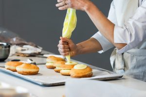 Side view of crop anonymous baker squeezing cream from pastry bag while decorating sweet vegan Berliners in kitchen in bakery