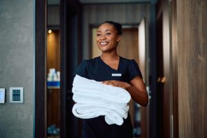 Happy African American housekeeper with stack of fresh towels in hotel room. Copy space.