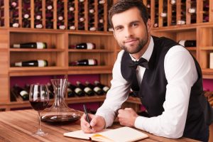 I know everything about wine. Confident male sommelier writing something in his note pad and looking at camera while leaning at the wooden table with wine shelf in the background